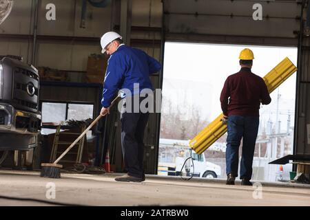 Man sweeping workshop while other carrying supplies Stock Photo