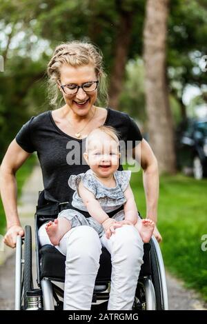 A paraplegic mom carrying her baby in her lap while using a wheelchair outdoors on a warm summer afternoon: Edmonton, Alberta, Canada Stock Photo