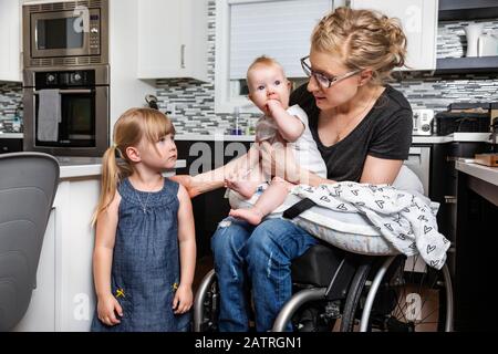 A paraplegic mom in a wheelchair talking with her daughter and holding her baby in her lap while working in her kitchen; Edmonton, Alberta, Canada Stock Photo