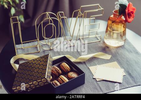 Festive box with chocolate macaroons cakes, rose flower in vase, postcard and wire word Love on gray napkin. Cozy gift idea for lover. Hygge Valentine Stock Photo