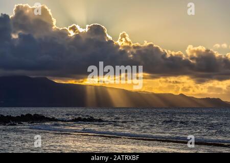 Beautiful sunlight rays stream through the dark clouds and fading daylight over the pacific ocean on a secluded Maui, Hawaii beach. The rocks and i... Stock Photo