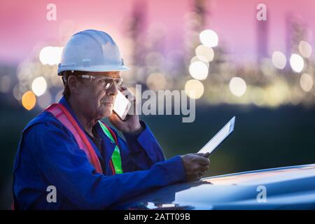 Engineer working on a tablet and smart phone with an oil refinery in the background; Alberta, Canada Stock Photo