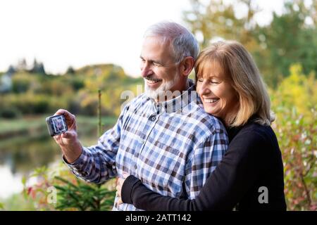 A mature couple enjoying time together and taking a self-portrait while enjoying the sunset along a river in a city park on a warm fall evening Stock Photo