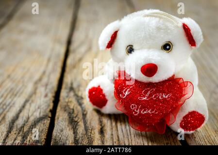 Cute white teddy bear holding a red heart and sits on an old wooden table, holiday gift for Valentine's Day. Stock Photo