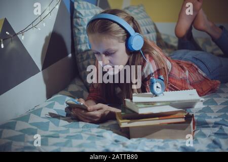 child and gadget addiction. girl in headphones in the childrens room lies on the bed and looks at the phone Stock Photo