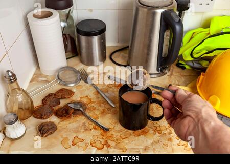 Builder making a cup of tea in his dirty kitchenette next to his hard hat and high visibility vest Stock Photo