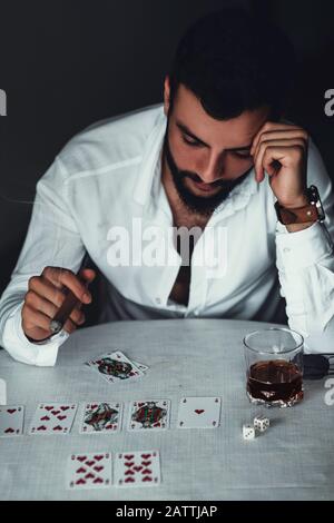 The young gambler looks troubled at the gambling table with whiskey and a cigarette in his hand. Gambling Concept. Stock Photo