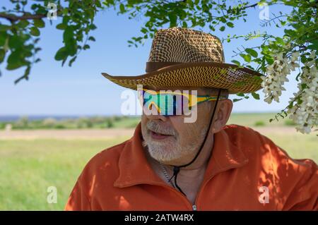 Portrait of bearded Caucasian senior farmer wearing chameleon sunglasses and standing under acacia tree against green agricultural field Stock Photo
