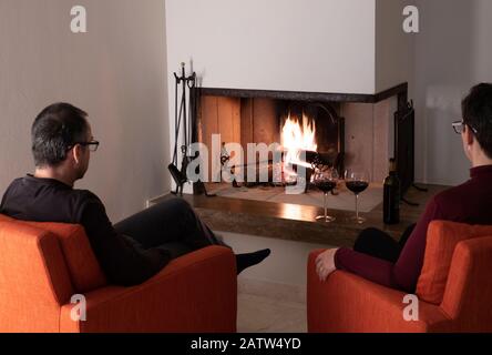 A middle aged couple enjoying a glass of red wine together in comfortable armchairs seated in front of a roaring fire in a fireplace viewed from the r Stock Photo