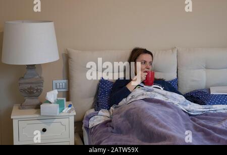 Sick woman sitting up against the pillows drinking a hot beverage in bed from a red mug and looking away to the side