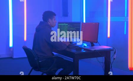 Young african man writing hacker programs in dark room with neon lights. He working on computer with two monitors. Stock Photo