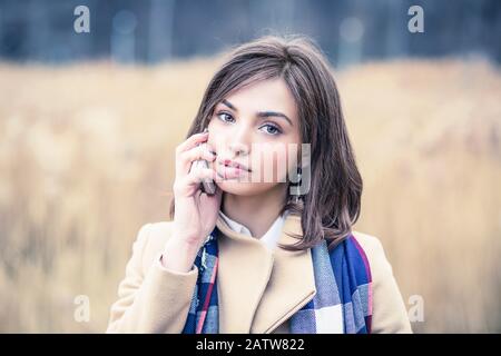 Woman girl talking on a mobile phone playing with her lips looking at you camera isolated outdoors light brown park background in autumn nature, weari Stock Photo