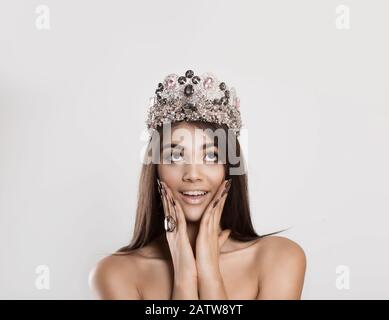 Wow, look up here. Happy beauty queen indian woman with crown looks up excited surprised, hands on her cheeks, against a white gray background Stock Photo