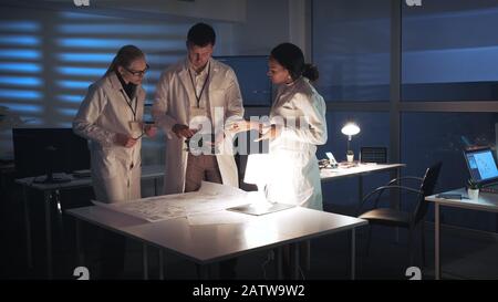 Technology Research Facility: Multiracial Electronics Development Engineers in white coats working in lab with motherboard and control electronics scheme. They discussing and analyzing details Stock Photo