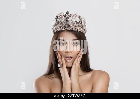 Wow, I won. Closeup young woman beautiful girl crowned miss long dark hair looking excited holding her mouth opened hands on cheeks white grey wall. S