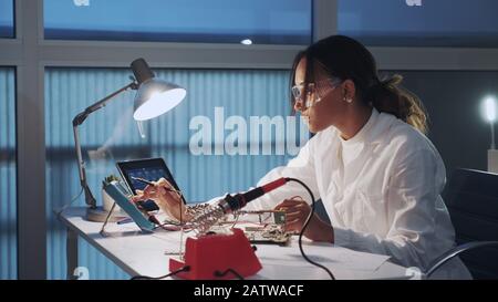 Close up of female electronics engineer in protective glasses checking motherboard with multimeter tester in advanced technology laboratory Stock Photo