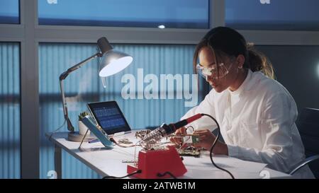 Close up of female electronics engineer in protective glasses checking motherboard with multimeter tester in advanced technology laboratory Stock Photo