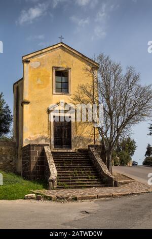 Yellow facade of a small church on the corner of the streets, surronded by trees and bushes. Staris to the entrance door. Cross on the roof. Sant´Orso Stock Photo