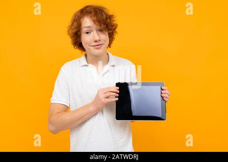 red-haired curly guy in a white T-shirt shows a tablet display with a layout on a yellow background Stock Photo