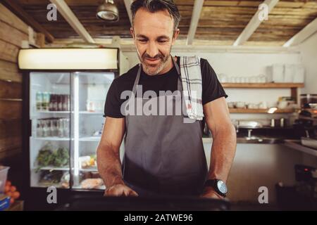 Male barista using cashbox computer at cafe counter for checking client's order. Mature man in apron working on a coffee shop computer. Stock Photo