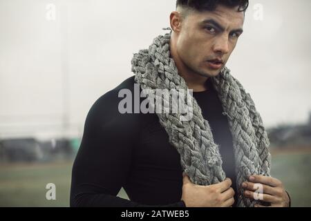 Muscular man relaxing after workout with a battle rope in his neck. Strong man taking rest after doing fitness workout outdoors. Stock Photo