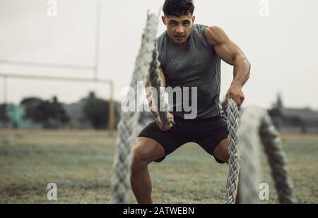 Muscular young man working out with battling ropes. Fit young male athlete doing battle rope workout outdoors on a field. Stock Photo
