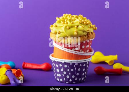 Birthday cupcake in funny polka dot colorful cups stack and deflated balloons on violet background, close up, copy space Stock Photo