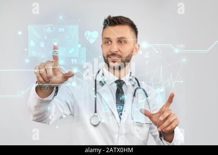 Front view of smiling doctor with strethoscope wearing lab coat. Selective focus of digital tactile charts screen, man touching virtual icons on projection. Concept of digitalization, medicine. Stock Photo