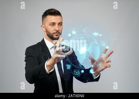 Front view of young serious businessman in black suit working in office. Man holding virtual 3D projection of digital tactile charts screen in arms. Concept of high technologies, digitalization. Stock Photo