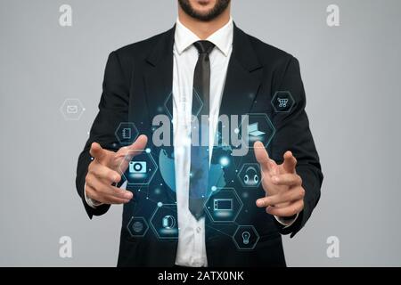 Front view of incognito businessman in black suit in office. Man holding virtual 3D projection of digital tactile charts screen in arms. Concept of high technologies, digitalization. Stock Photo
