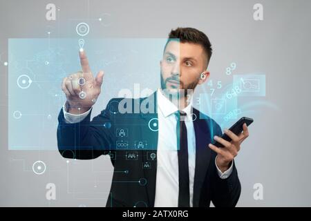 Front view of businessman in suit holding phone with virtual projection in office. Serious bearded man clicking virtual button on digital tactile charts screen. Concept of gadgets, digitalization. Stock Photo
