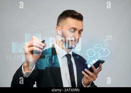 Front view of businessman in suit holding phone with virtual projection in office. Serious man clicking virtual button on digital tactile charts screen with stylus. Concept of gadgets, digitalization. Stock Photo