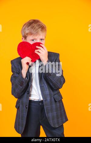 Little blond boy holding heart and laughing at camera on yellow background. Stock Photo