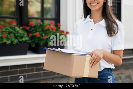 Crop of incognito happy joyful young female courier in white blouse and jeans carrying box and folder. Brunette woman standing outdoors, holding parcel and smiling. Delivery concept. Stock Photo