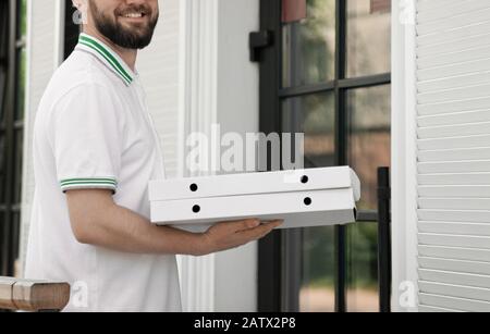 Crop of incognito happy joyful young male courier in white polo carrying pizza outdoors. Bearded man standing near door, holding two pizza boxes and smiling. Food delivery concept. Stock Photo