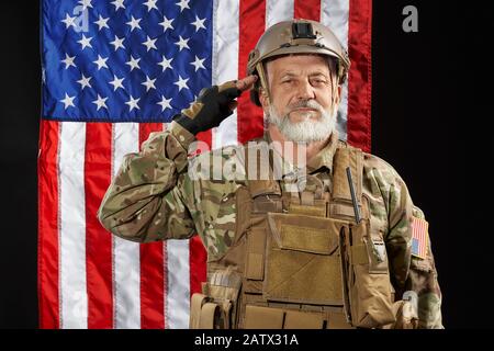 Front view of old military officer saluting and looking at camera. Portrait of bearded american veteran in uniform and helmet posing on black background with flag. Concept of military, army. Stock Photo