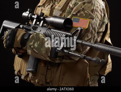 Front view of incognito military officer holding sniper rifle. Crop of american soldier in uniform and helmet posing with gun on black studio background. Concept of army, military. Stock Photo