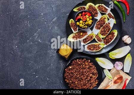 close-up of Endive Boats with beef ragout and chopped green olives fillings on a black plate with ingredients on the background, horizontal view from Stock Photo
