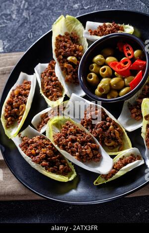 low-carb Ground Beef and chopped green olives Endive Boats served on a black plate on a grey concrete table, vertical view from above, close-up Stock Photo