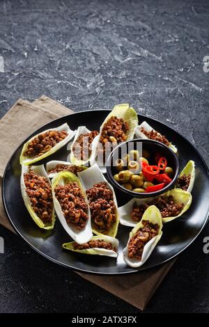 low-carb Ground Beef and chopped green olives Endive Boats served on a black plate on a grey concrete table, vertical view from above, free space Stock Photo