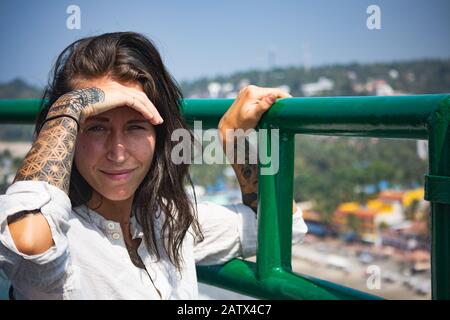 Beautiful girl with many tattoos on both arms shields her face from the strong sunlight whilst looking at the camera with a great view Stock Photo