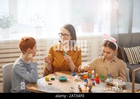 Mother with her two children sitting at the table and decorating eggs together for Easter Stock Photo