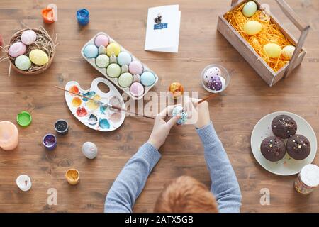 High angle view of boy sitting at wooden table with cakes and eggs and decorating them with colorful paints Stock Photo