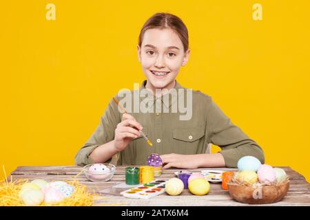 Portrait of happy girl sitting at the table and smiling at camera while painting Easter eggs against the yellow background Stock Photo