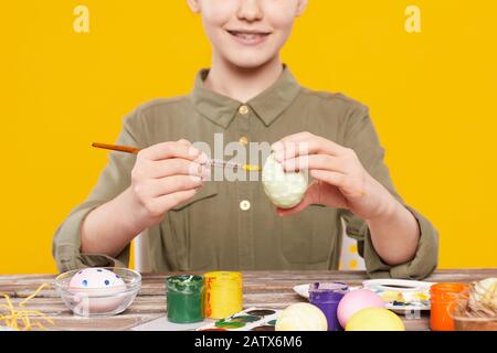 Close-up of smiling girl sitting at the table and decorating eggs with multicolored paints for Easter against the yellow background Stock Photo
