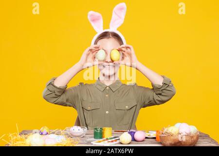 Portrait of funny girl in rabbit ears holding Easter eggs in front of her face and smiling while sitting at the table with paints Stock Photo