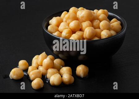 Cooked chick peas in a black ceramic bowl isolated on black painted wood. Spilled chick peas. Stock Photo