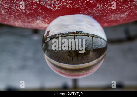 Remote port in lensball, focus on foreground, wooden footbridge with railing. Abstract representation of reality by looking into a magic sphere. Lake Stock Photo