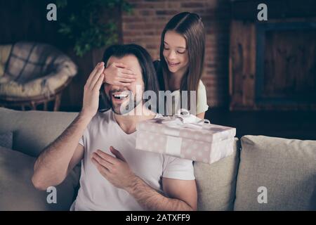 Guess who. Photo of little adorable pretty lady her handsome young daddy sit cozy sofa hiding dad eyes holding big surprise giftbox domestic homey Stock Photo