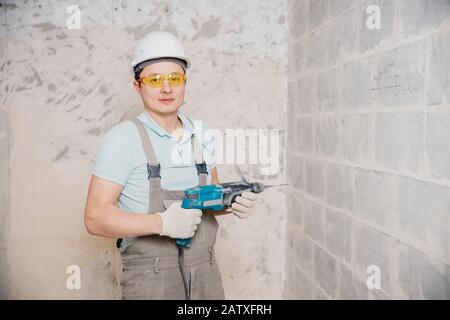 Portrait man builder repairer in protective glasses, hard hat, overalls showing having equipment in arm on grey background home brick Stock Photo